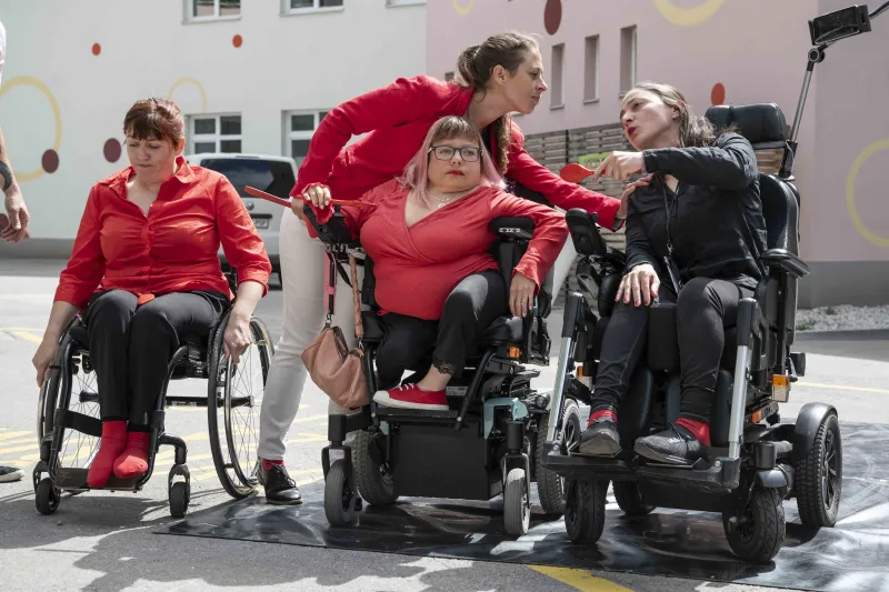 Na fotografiji so gibalno ovirane osebe na treh invalidskih vozičkih. Ob njih je spremljevalna oseba. Oblečeni so v rdeča in črna oblačila. Med seboj komunicirajo.