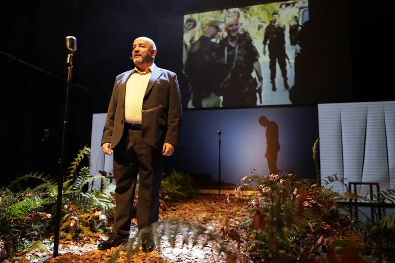 Moški pred mikrofnom, po tleh listje in okrog njega praprot; za njim na platnu videoposnetki vojakov
