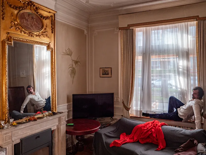 Moški sedi na okenski polici in gleda proti ogledalu na steni, pod njim je razmetan kavč in v kotu televizija.